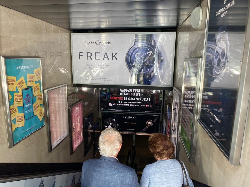 Campagne publicitaire lumineuse déployée au parking du Mont-Blanc à Genève : affiches rétroéclairées positionnées en fronton et en latéral autour des escalators, assurant une visibilité totale des flux piétons.