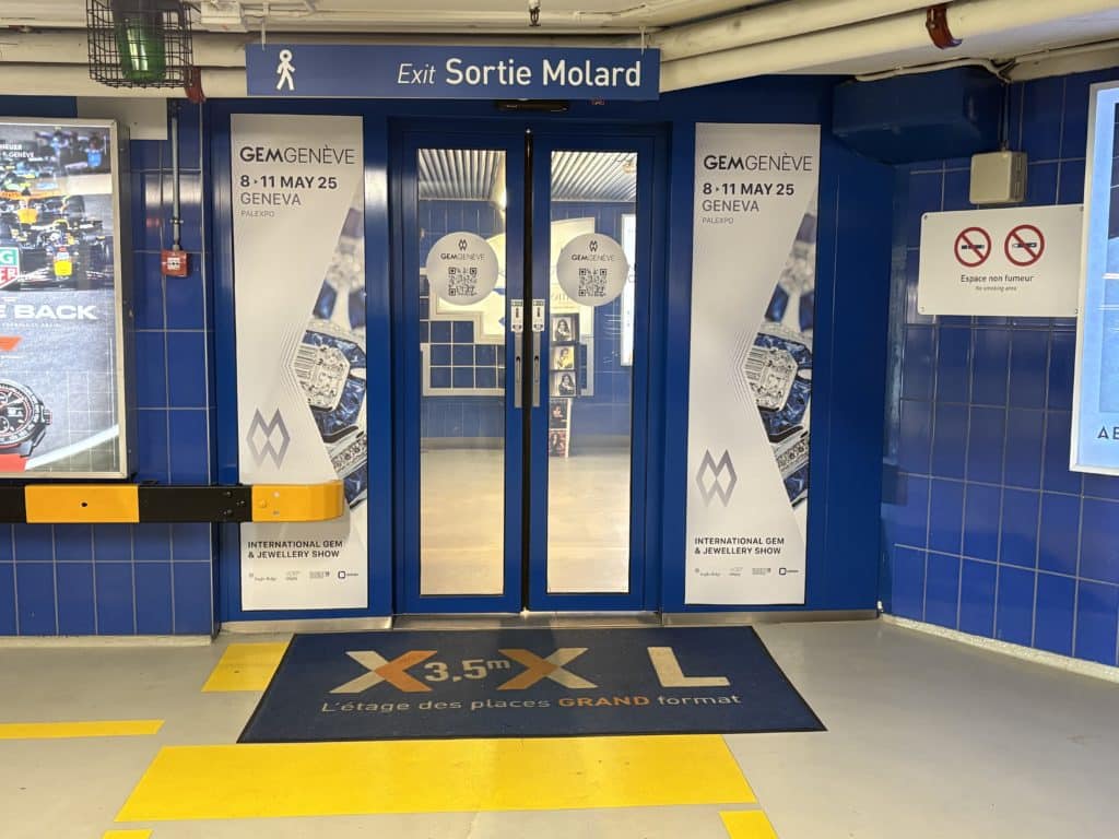 Visuel événementiel GemGenève installé sur les portes coulissantes automatiques du parking du Mont-Blanc à Genève, touchant un public haut de gamme en transit.