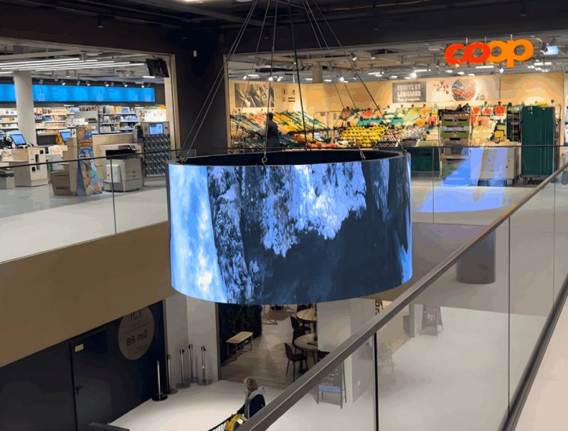 Rotating 360° digital screen displaying an advertisement at M Central shopping center in Monthey, visible from all angles to visitors.