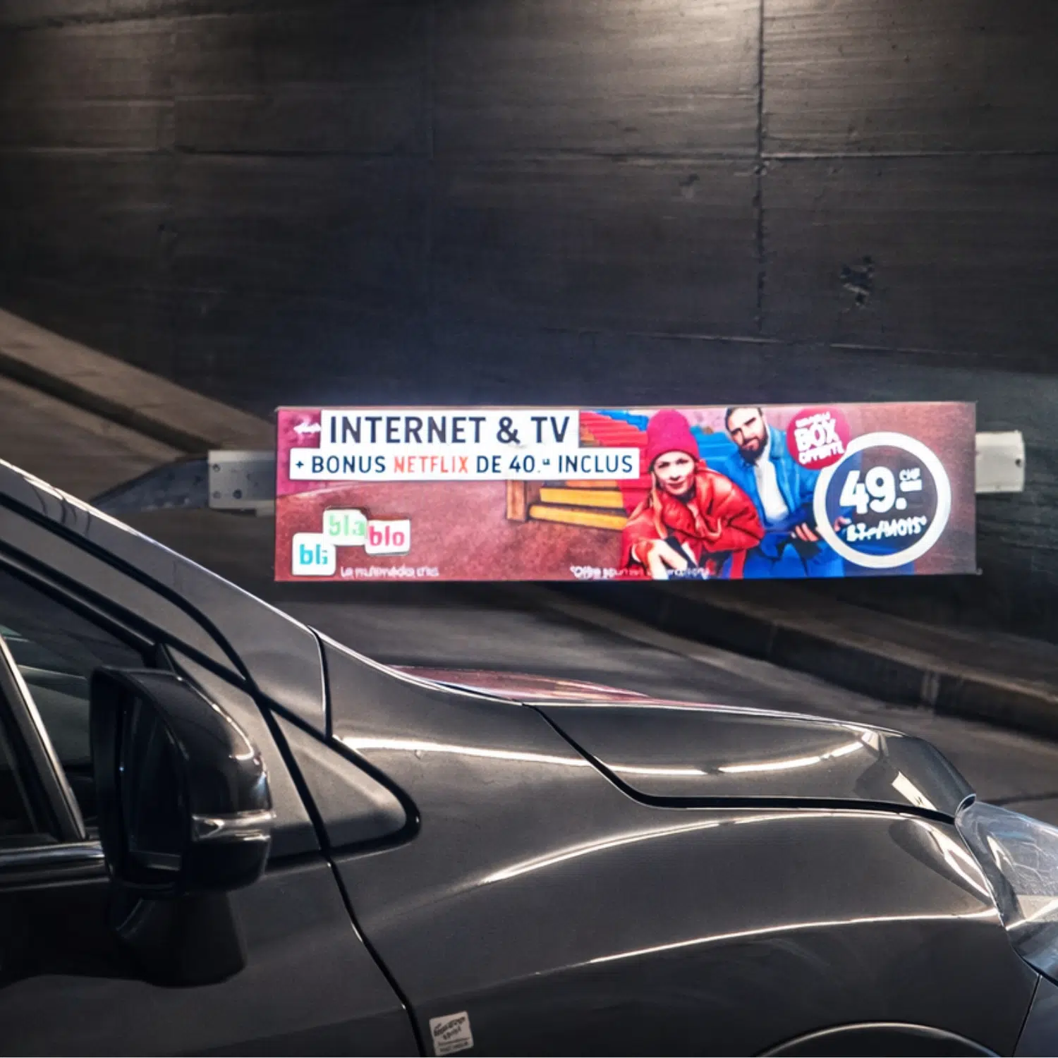 Barrière publicitaire BliBlaBlo pour une offre Internet et TV avec bonus Netflix, installé dans le parking souterrain du centre Manor à Sion, avec une voiture au premier plan.