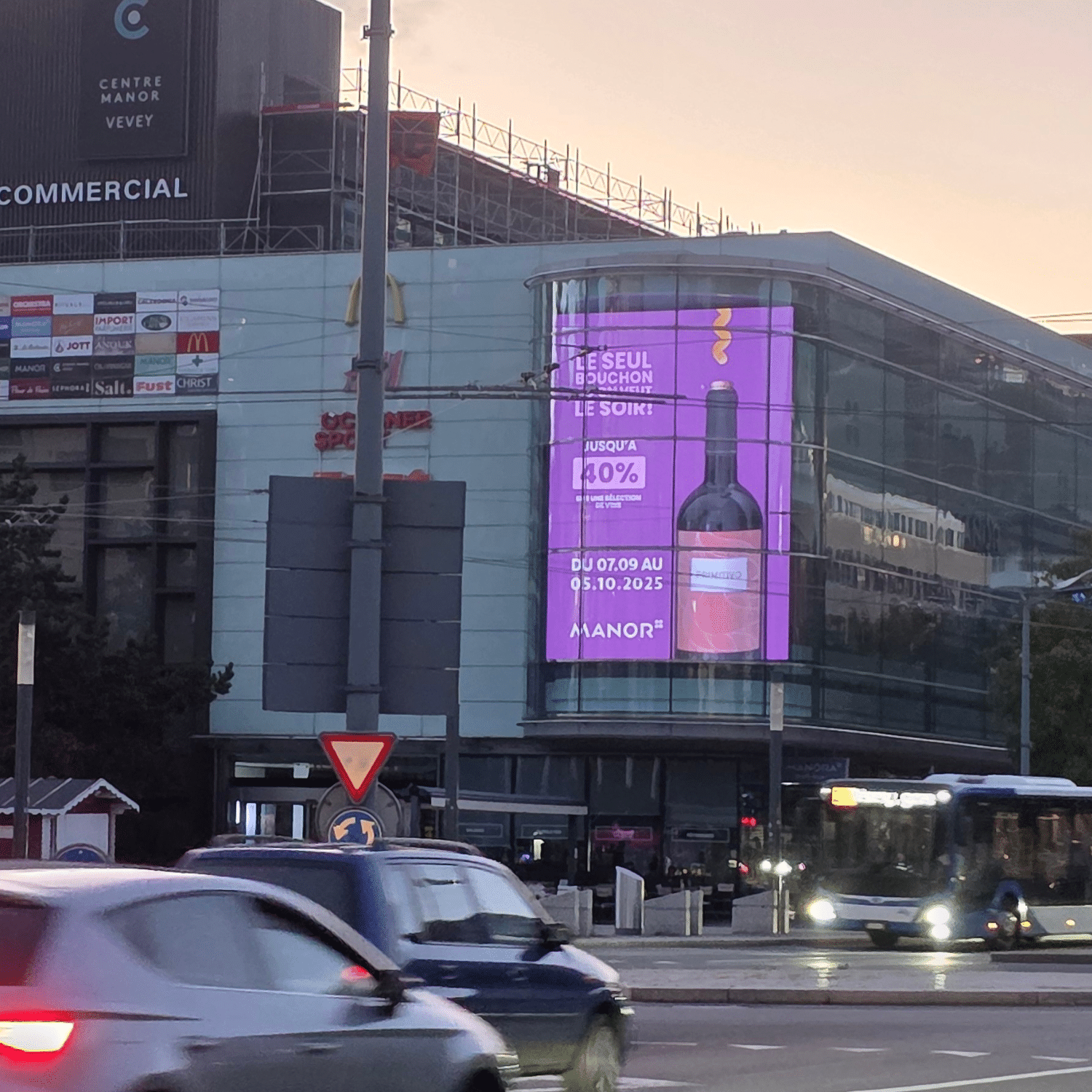 Grand mégaposter affiché sur la façade du magasin Manor au centre-ville de Vevey, visible depuis la rue et offrant une exposition maximale aux passants.