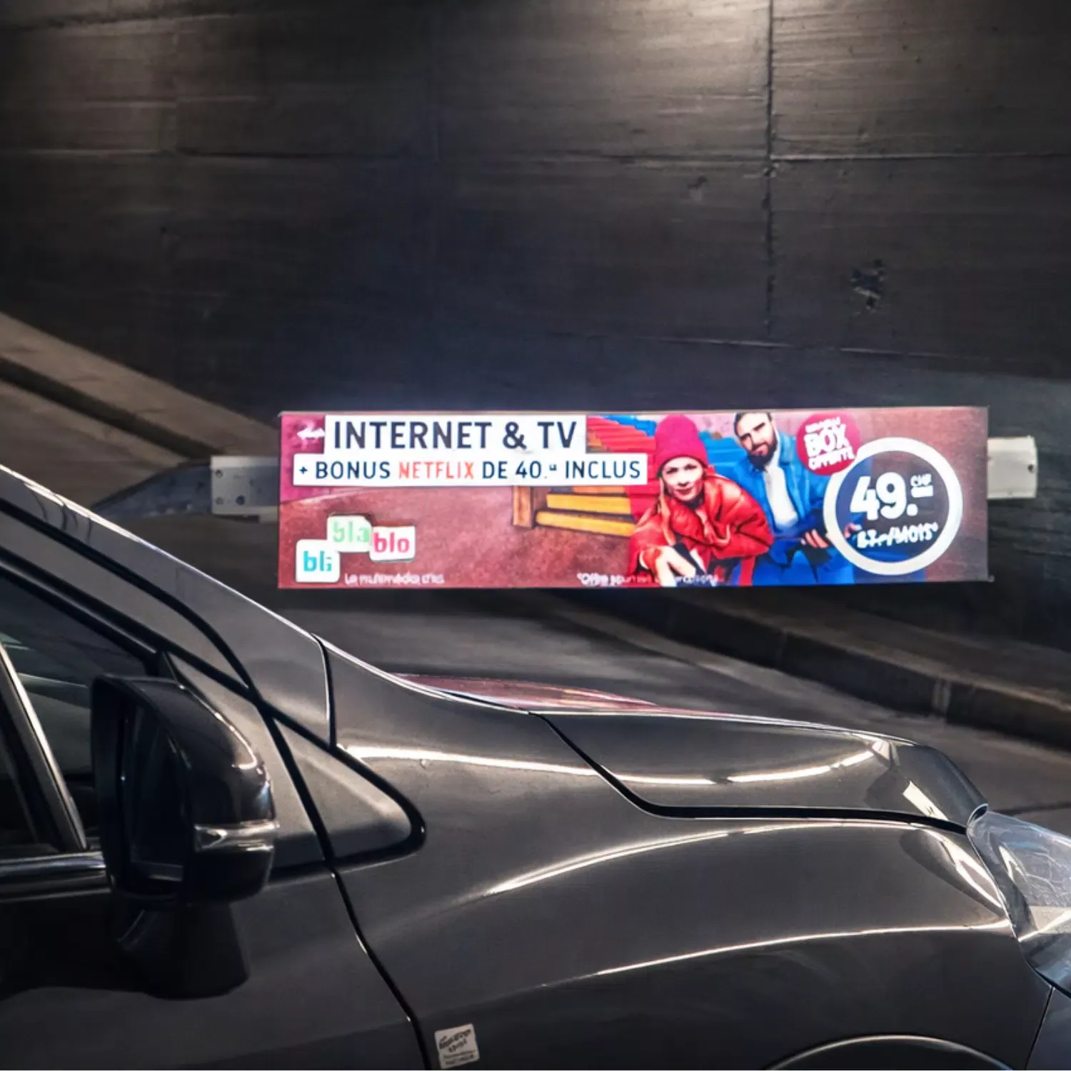Barrière publicitaire BliBlaBlo pour une offre Internet et TV avec bonus Netflix, installé dans le parking souterrain du centre Manor à Sion, avec une voiture au premier plan.