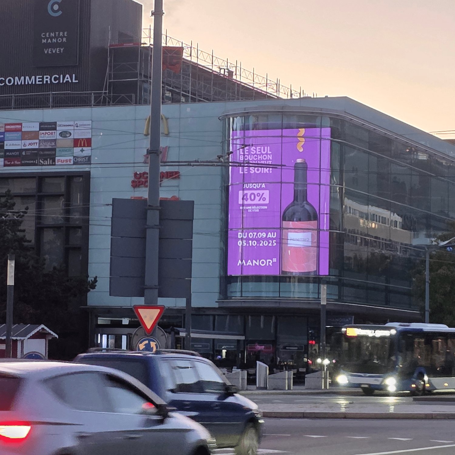 Grand mégaposter affiché sur la façade du magasin Manor au centre-ville de Vevey, visible depuis la rue et offrant une exposition maximale aux passants.