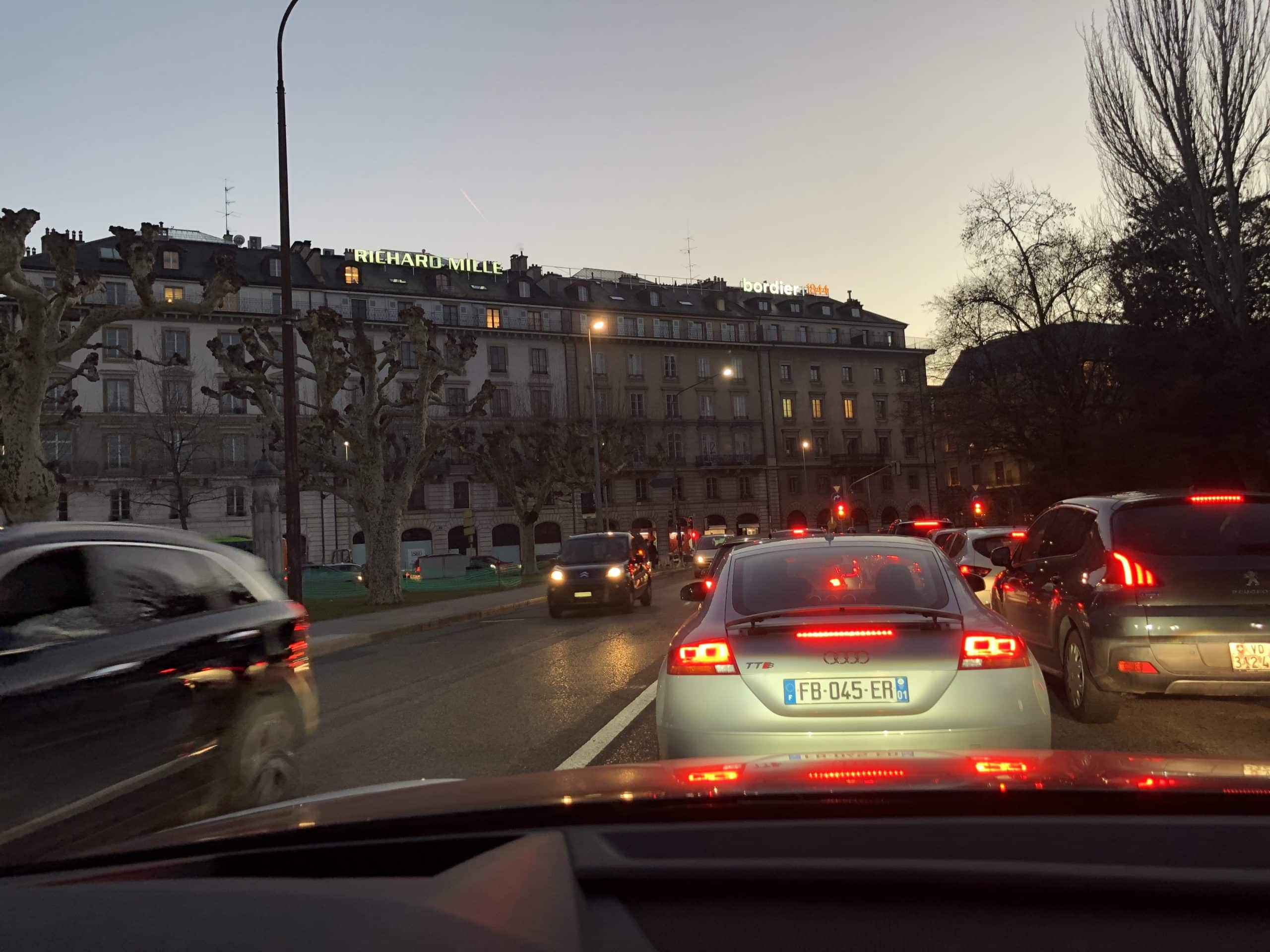 Night photograph of the Richard Mille illuminated sign on the façade of an elegant building on Quai Général-Guisan in Geneva, with reflections of the lights on the street and a refined urban atmosphere.