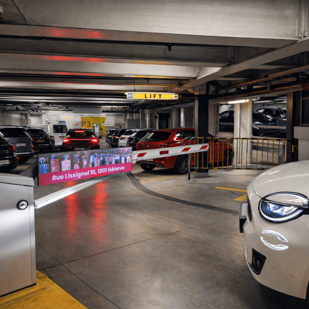 Barrière de parking équipée d’un écran LED diffusant la publicité Cuvées du Terroir dans le parking souterrain de Manor Genève, avec véhicules stationnés et signalétique intérieure visible.