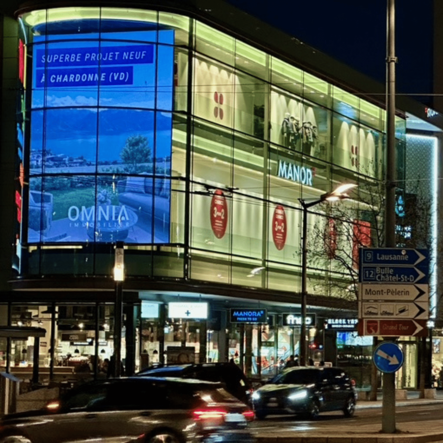 Grand écran publicitaire digital de 46 m² situé à Vevey, diffusant une campagne Omnia Immobilier sur la façade vitrée d’un centre commercial en environnement urbain nocturne avec circulation et éclairage dynamique.