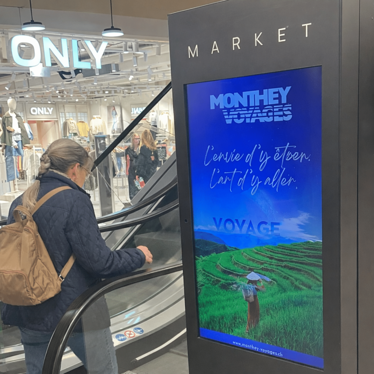 Une borne publicitaire digitale grand format, installée à proximité immédiate des escalators du centre commercial MCentral à Monthey, diffuse une campagne Monthey Voyages. L’écran lumineux capte l’attention des flux de visiteurs en mouvement, offrant une visibilité optimale dans une zone de fort passage. Le visuel immersif de rizières et le message d’évasion renforcent l’impact du support dans cet environnement dynamique.