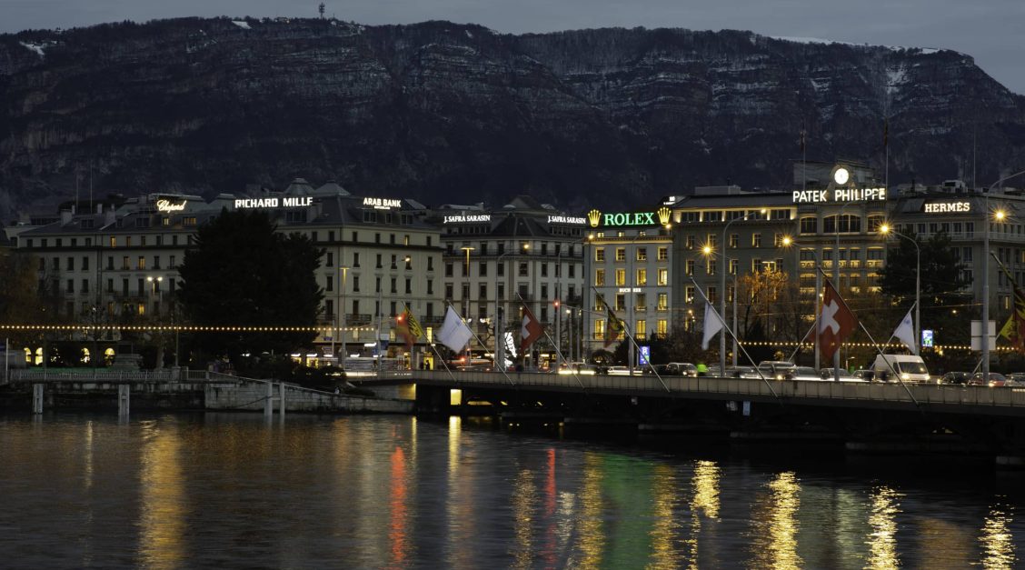 Night view of the Mont Blanc bridge in Geneva, with the illuminated signs on the roofs visible in the background, a symbol of prestige advertising.